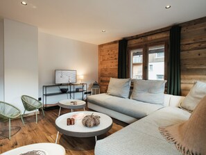 Ein modernes Wohnzimmer mit einer Couch, zwei Sesseln, einem runden Couchtisch, einem Fernseher auf einem Schrank und einer Holzvertäfelung.