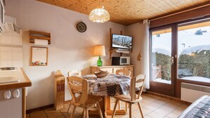 Ein gemütliches Zimmer mit Holzbalkendecke, einem Esstisch mit Stühlen, einem Fernseher und Blick auf eine verschneite Landschaft.