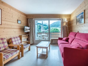 A cozy room with wooden walls, a red sofa, a wooden bench with plaid cushions, and a table. A large window offers a scenic view of a mountain and a church.