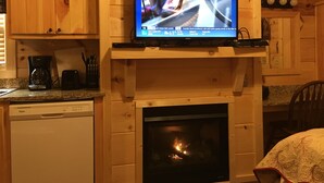 Un intérieur en bois avec une cheminée, un téléviseur fixé au-dessus, et un coin cuisine équipé d'une machine à café et d'ustensiles.