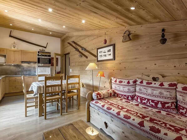 Une pièce chaleureuse avec un plafond en bois, un lit recouvert d’une literie à motifs, une table à manger avec des chaises, et un coin cuisine doté de meubles et d’appareils électroménagers en bois.