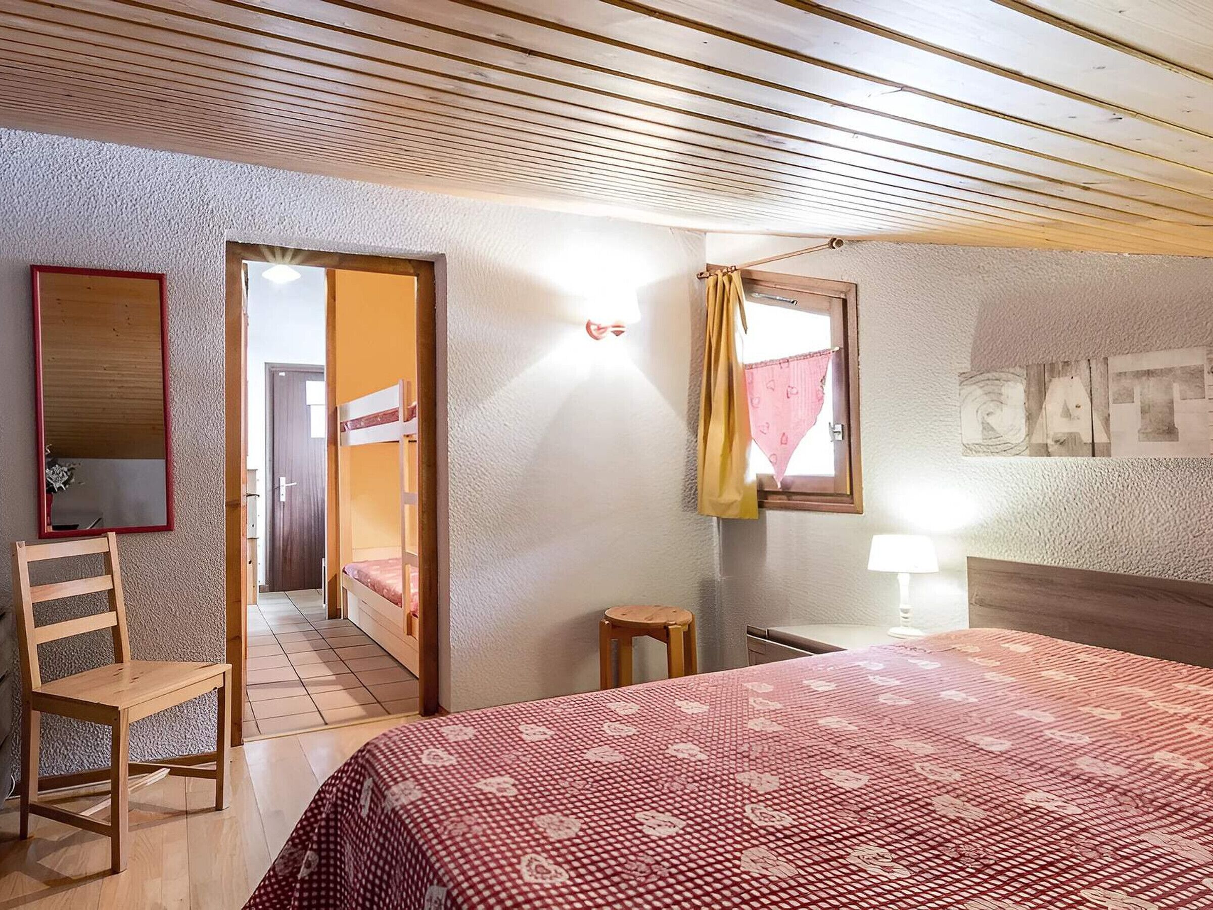 A bedroom with a bed, a chair, a small table, a mirror, and a door leading to another room.