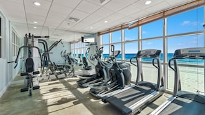A gym with treadmills, elliptical machines, and weight equipment. Large windows offer a view of the beach. - NEWLY UPDATED FURNITURE! | Outdoor Pool | Hot Tub | Sauna | BEACHBALL PROPERTIES (Gulf Shores)