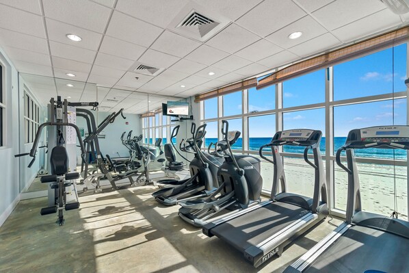 A gym with treadmills, elliptical machines, and weight equipment. Large windows offer a view of the beach. - NEWLY UPDATED FURNITURE! | Outdoor Pool | Hot Tub | Sauna | BEACHBALL PROPERTIES (Gulf Shores)