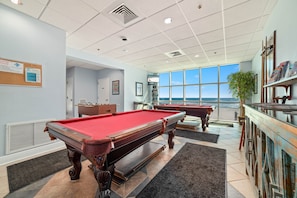 A pool table in a room with large windows, a foosball table, and a bookshelf. - NEWLY UPDATED FURNITURE! | Outdoor Pool | Hot Tub | Sauna | BEACHBALL PROPERTIES (Gulf Shores)