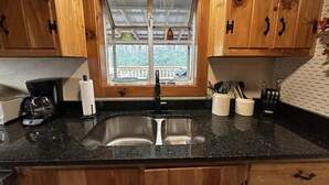 Una cocina con armarios de madera, encimera de granito negro, pileta de acero inoxidable y ventana con vista a vegetación.