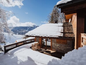 A wooden chalet with a stone foundation, a covered patio with seating, and a view of snow-covered mountains.