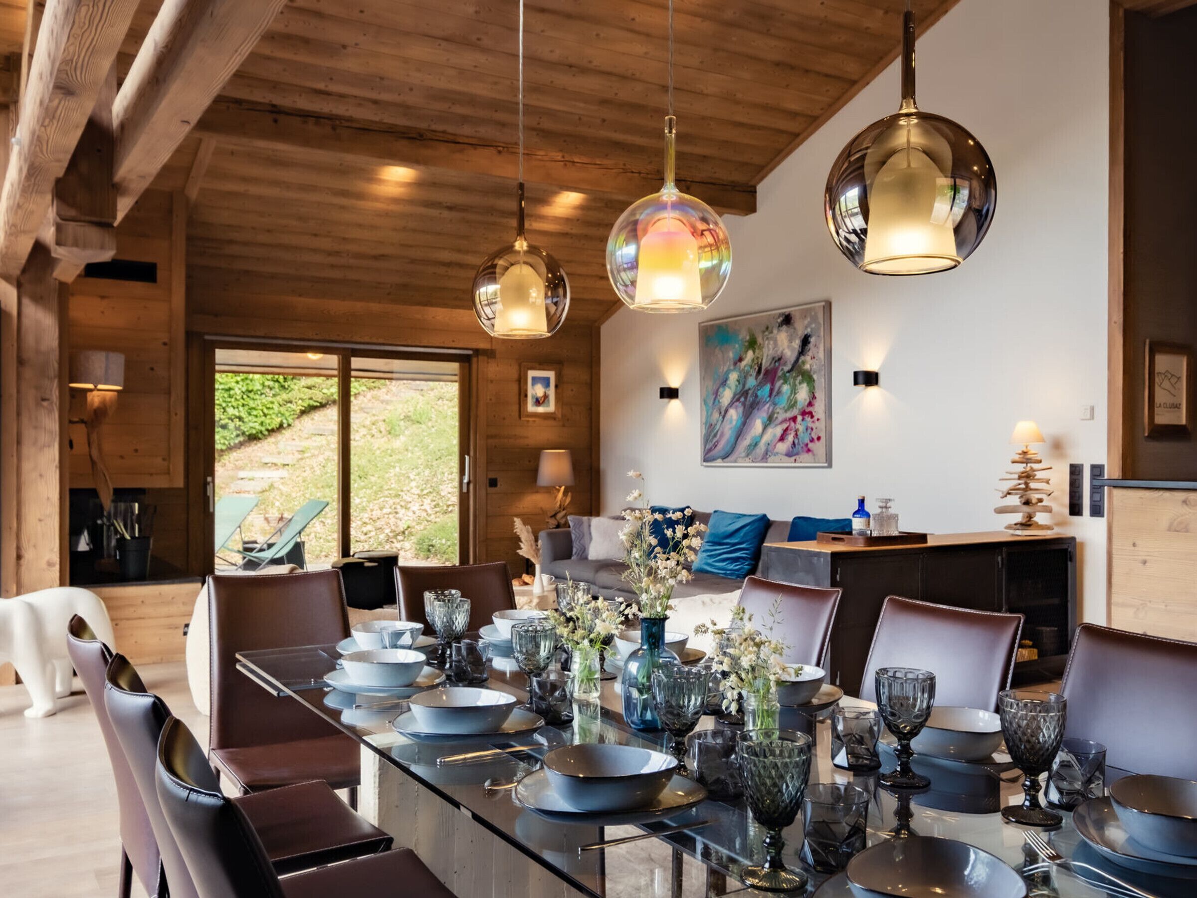 A modern dining area with a long table set for a meal, wooden ceiling beams, and a painting on the wall.