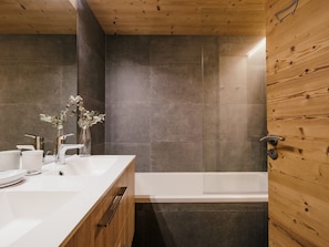 A modern bathroom with a wooden ceiling, a white sink, a glass shower enclosure, and a dark tiled wall.