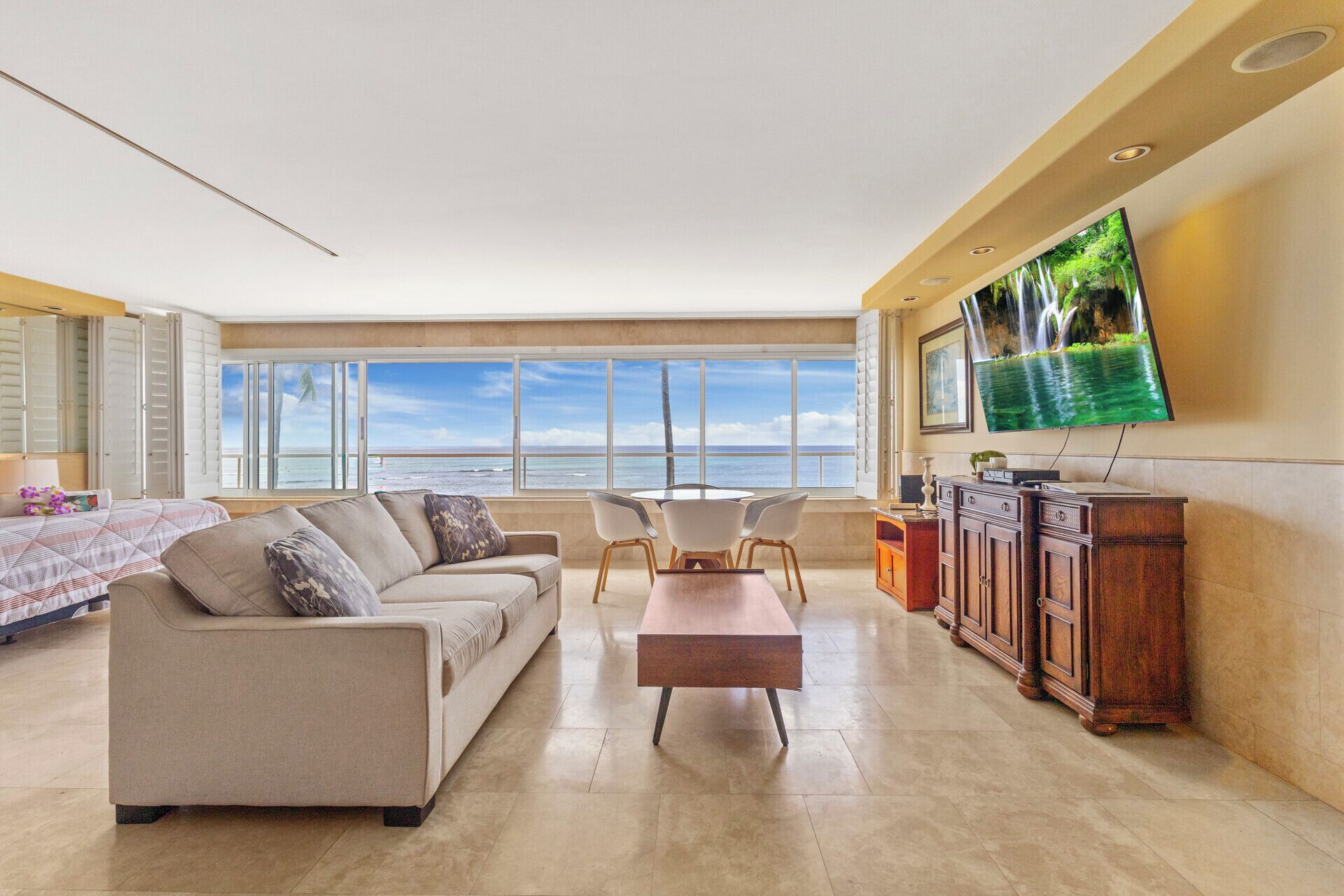 A spacious living room with a large window overlooking the ocean, a comfortable sofa, a dining table with chairs, and a flat-screen TV mounted on the wall.