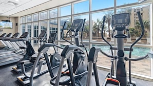 Un gimnasio con cintas de correr y equipamiento de ejercicio, amplias ventanas y vista a una piscina y palmeras.