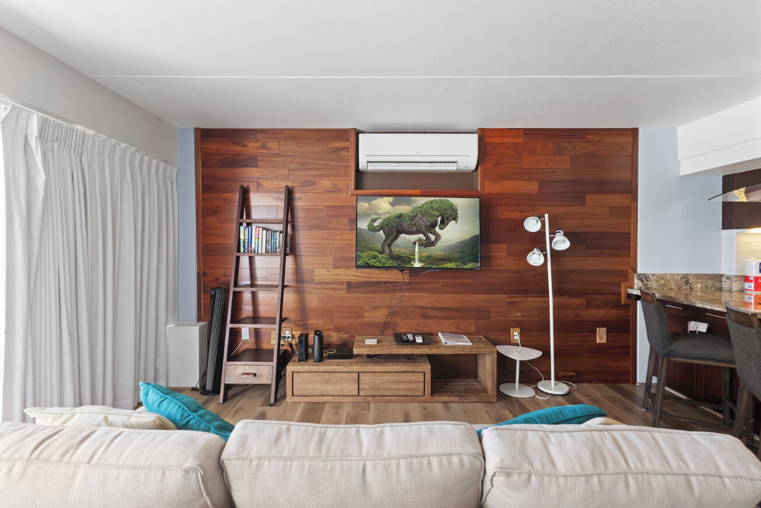 A modern living room with a wooden wall, a mounted TV, a bookshelf, a sofa, and a dining area with a bar.