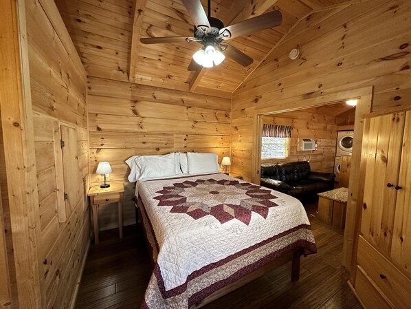 A wooden cabin bedroom with a bed, two bedside tables with lamps, a ceiling fan, and a washing machine in the corner. - Get N' Lucky – Cozy Mountain Cabin w/ Hot Tub & Views (Asheville)