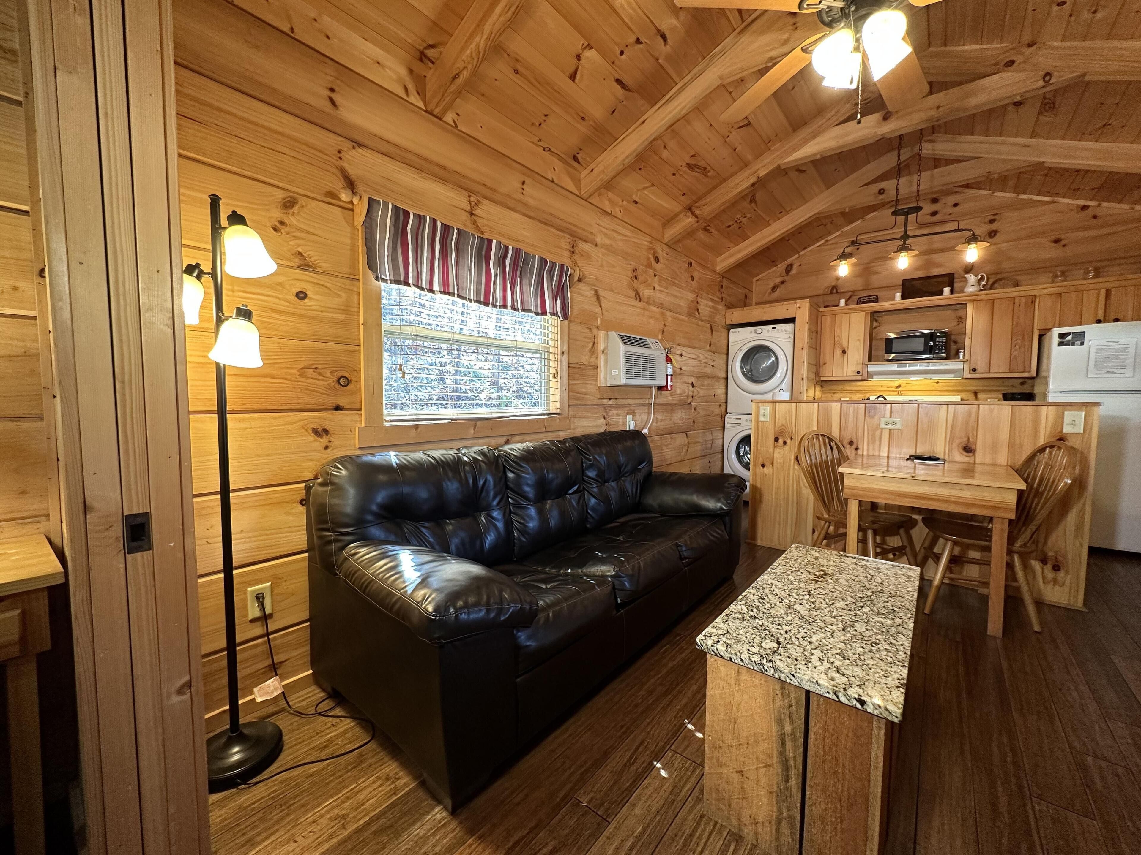Un acogedor interior de cabaña con un sofá de cuero negro, una mesa de comedor de madera con sillas, una zona de cocina con electrodomésticos y una ventana con cortinas.