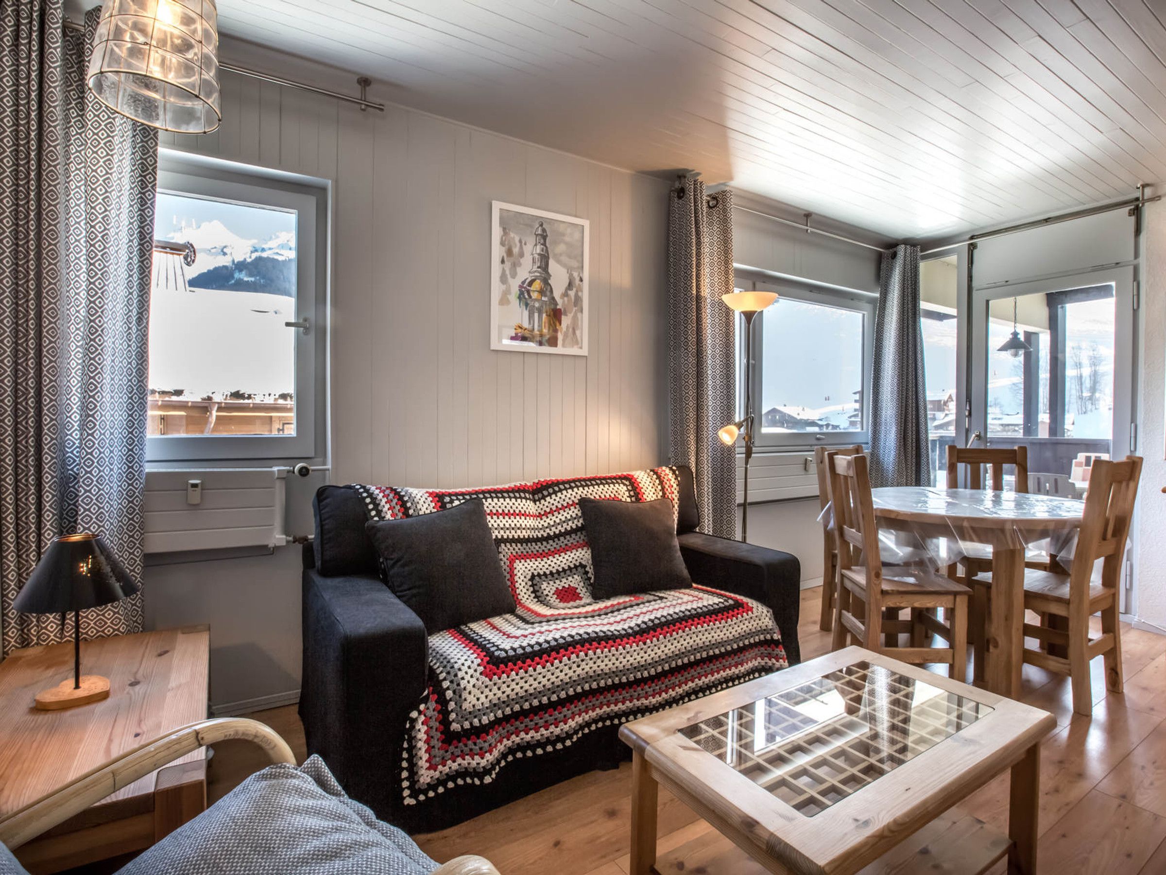 A cozy living room with a sofa, a coffee table, and a dining table with chairs. There is a large window with a view of mountains and a painting on the wall.