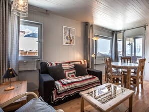Un salón acogedor con sofá, una mesa de centro y una mesa de comedor con sillas. Hay un ventanal con vistas a montañas y un cuadro en la pared.