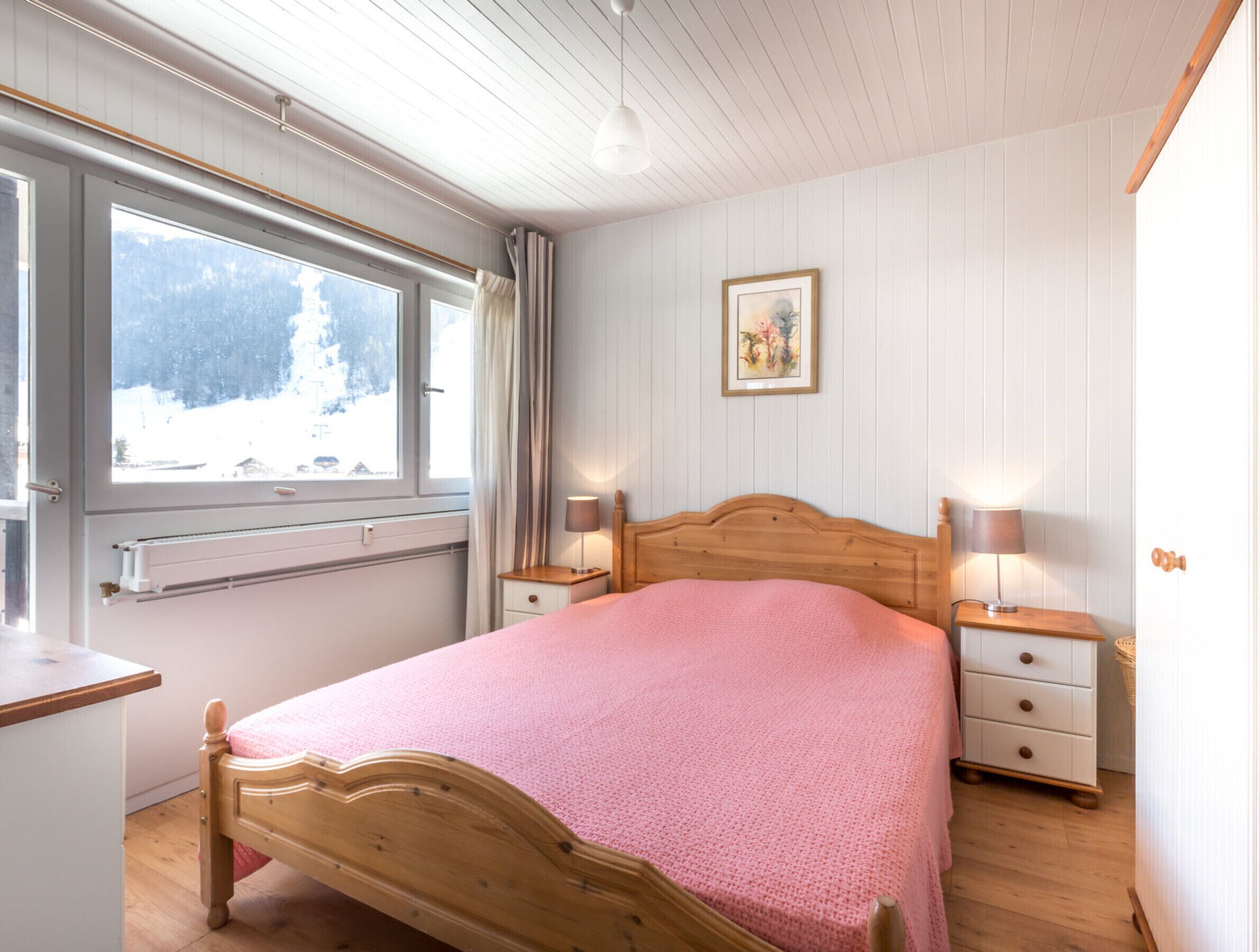 A bedroom with a wooden bed, a window with curtains, a bedside table with a lamp, and a wall-mounted picture.