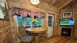 A wooden interior with a round wooden table and chairs, a fireplace, and a television displaying a weather forecast. - “Lovey Dovey: Your Romantic Asheville Cabin Escape” (Asheville)