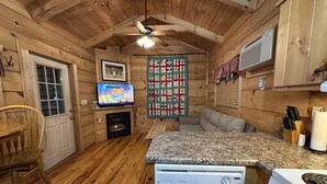 A wooden cabin interior with a kitchen area, a fireplace, a television, and a ceiling fan. - “Lovey Dovey: Your Romantic Asheville Cabin Escape” (Asheville)