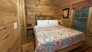 A wooden cabin bedroom with a bed, a nightstand, a lamp, a window with blinds, and a ceiling fan. - “Lovey Dovey: Your Romantic Asheville Cabin Escape” (Asheville)