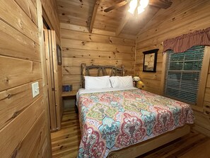 Quarto de cabana de madeira com cama, mesa de cabeceira, abajur, janela com persianas e ventilador de teto.