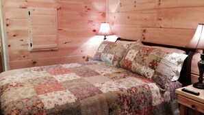 A wooden-paneled room with a bed, a nightstand, a lamp, and a window with blinds.