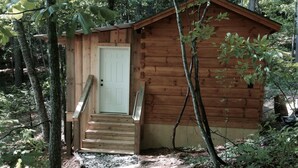 A wooden cabin with a white door and wooden steps leading up to it, surrounded by trees and a gravel path.