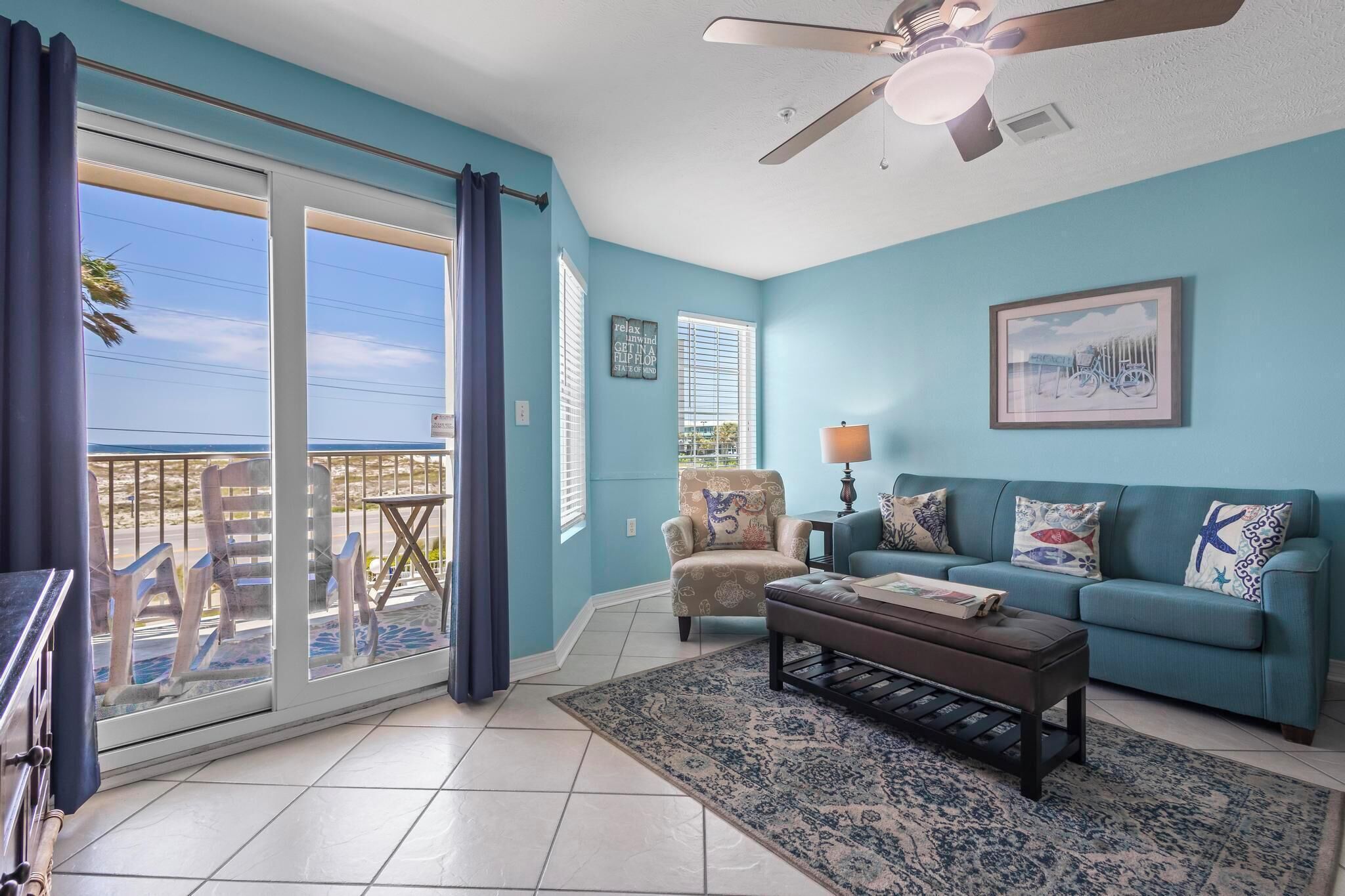 A living room with a teal wall, a ceiling fan, a sofa with patterned cushions, a coffee table, and a view of the outdoors through a sliding glass door.