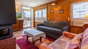 A cozy living room with a fireplace, wooden walls, a sofa, a table, and a view of the outdoors. - 3* Apartment in La Clusaz, 5 guests, parking (La Clusaz)