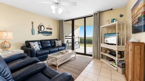 A living room with a sofa, coffee table, and TV, a ceiling fan, and a view through a glass door. - DIRECT GULF FRONT UNIT, CHARMING FIRST FLOOR LOCATION, TONS OF FAMILY AMENITIES, (Orange Beach)