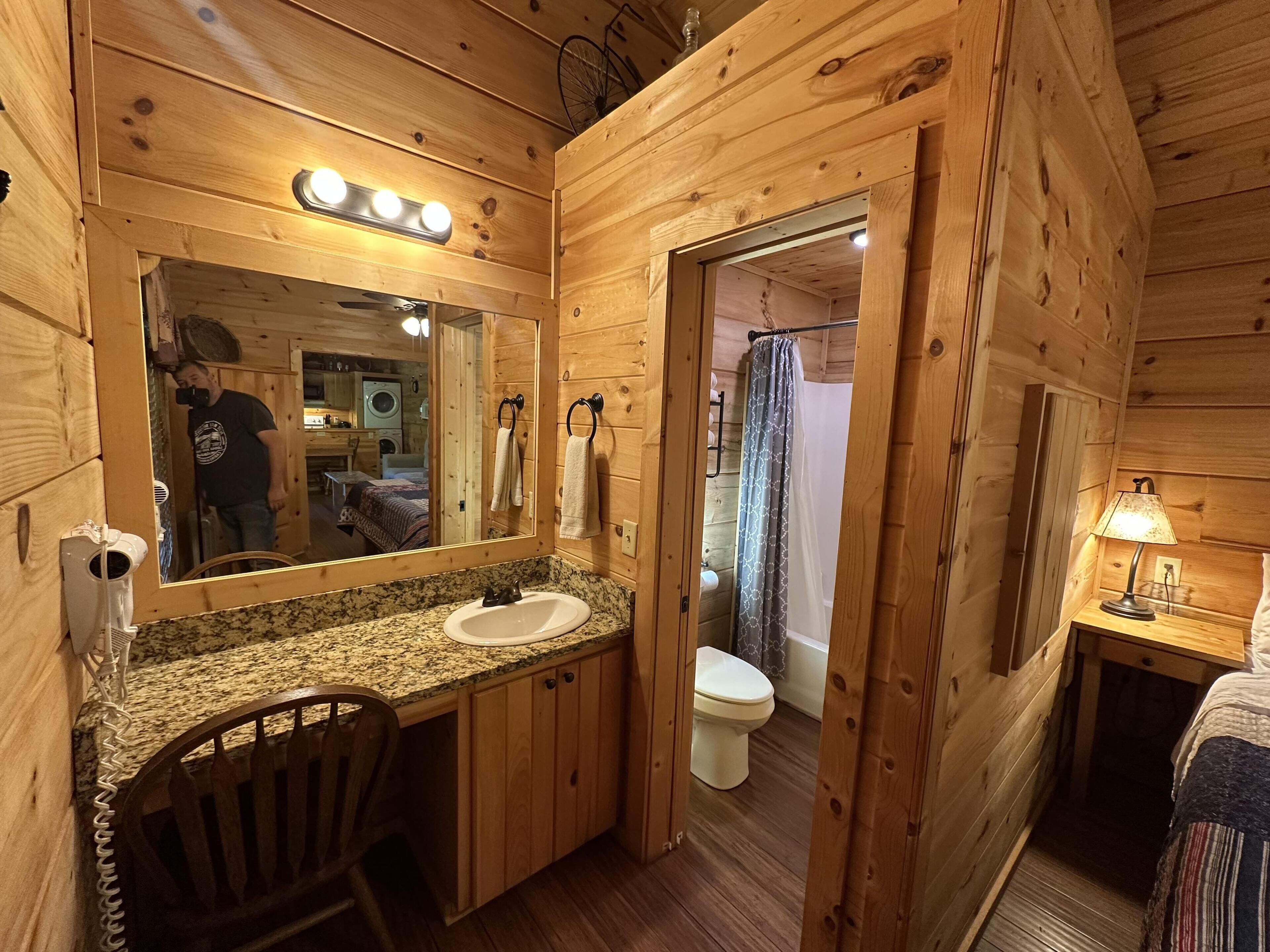 A wooden cabin bathroom with a granite countertop sink, a mirror, a towel rack, a shower curtain, and a bedside table with a lamp.