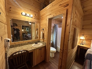 Une salle de bain rustique, avec une cabine en bois, un lavabo avec un plan de travail en granit, un miroir, un porte-serviettes, un rideau de douche et une table de chevet avec une lampe.