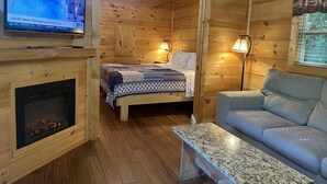 Ein Holzhütten-Interieur mit einem Fernseher, der Wetterinformationen anzeigt, einem Kamin, einem Bett mit einem gemusterten Teppich, einer Couch und einem Tisch.