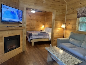 LâintĂ©rieur dâun chalet en bois comprend une tĂ©lĂ©vision qui affiche les prĂ©visions mĂ©tĂ©orologiques, une cheminĂ©e, un lit recouvert dâune couverture Ă motifs, un canapĂ© et une table.
