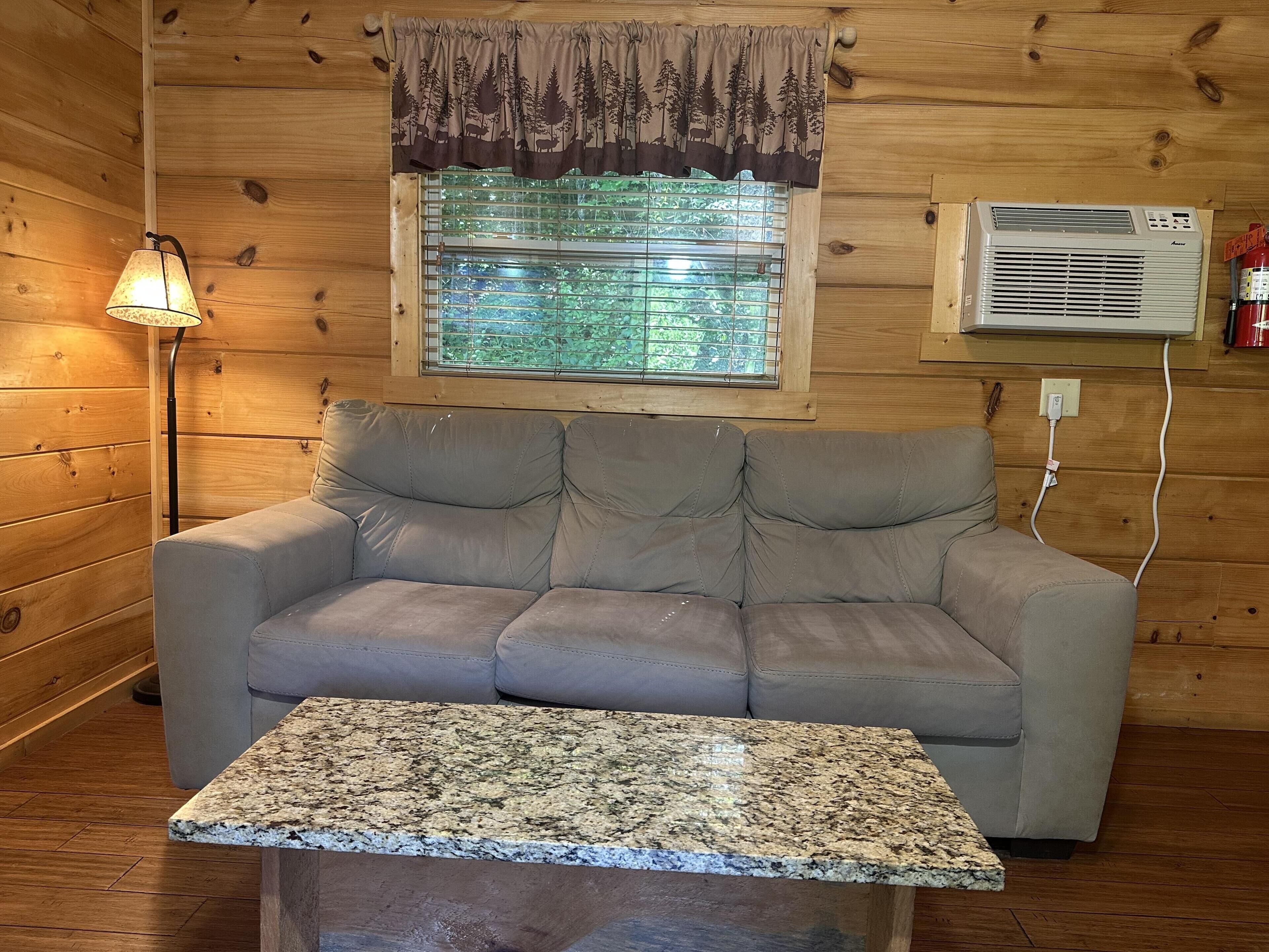 A wooden cabin interior with a sofa, a table, a window with a view of trees, and an air conditioning unit.