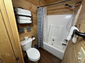 Une salle de bain aux murs lambrissĂ©s, Ă©quipĂ©e de toilettes, dâun porte-serviettes avec des serviettes pliĂ©es, dâun rideau de douche et dâune baignoire.