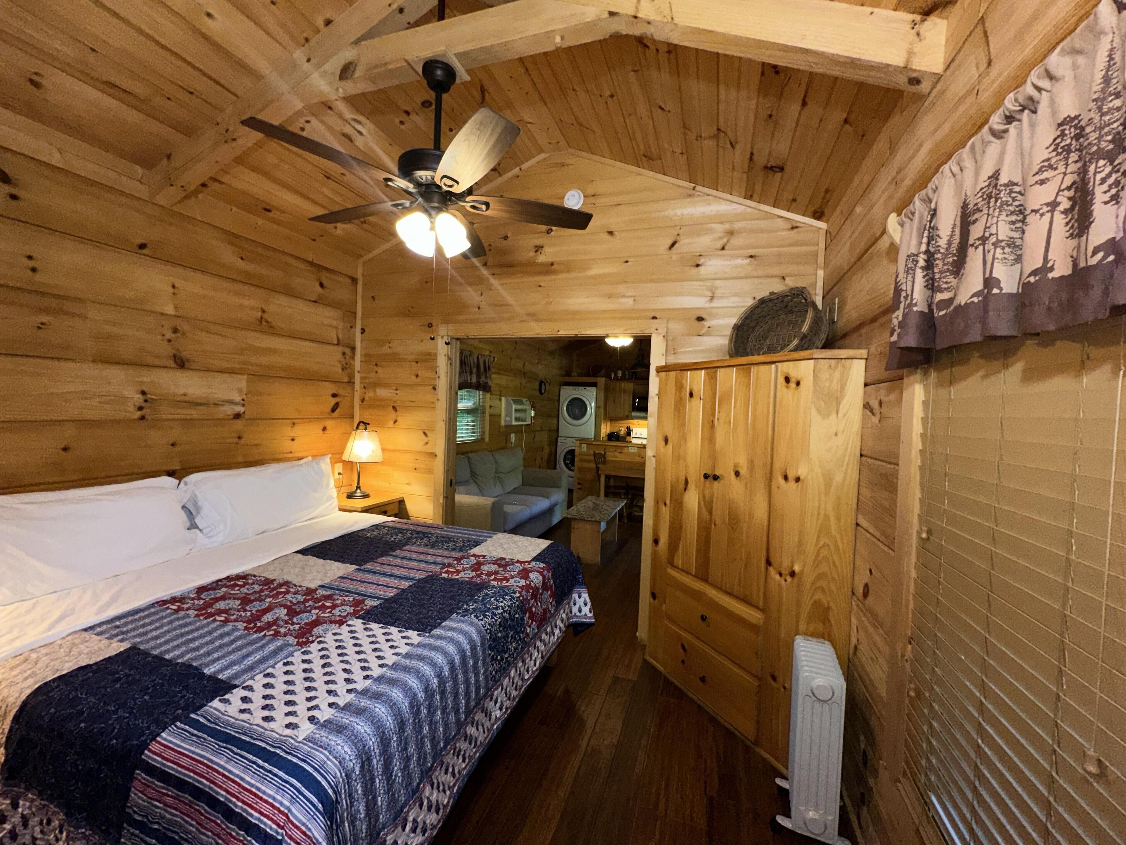 Habitació d'una cabana de fusta amb un llit, un ventilador de sostre, una tauleta de nit, una làmpada, un armari i vista a la cuina.