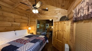 Ein Zimmer in einer Holzhütte mit einem Bett, einem Deckenventilator, einem Nachttisch, einer Lampe, einem Kleiderschrank und Blick in den Küchenbereich.