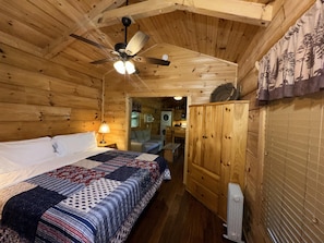 Une chambre dans une cabane en bois, comprenant un lit, un ventilateur de plafond, une table de chevet, une lampe, une armoire et une vue sur la cuisine.