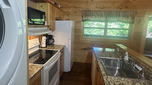 Eine kompakte Küche mit Holzausstattung, einer integrierten Mikrowelle, einem Herd, einem Kühlschrank, einer Spüle und einem Fenster mit Blick auf Grünflächen.