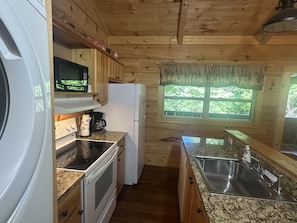Une cuisine compacte avec des meubles en bois, un four Ă micro-ondes encastrĂ©, une cuisiniĂšre, un rĂ©frigĂ©rateur, un Ă©vier et une fenĂȘtre donnant sur un espace verdoyant.