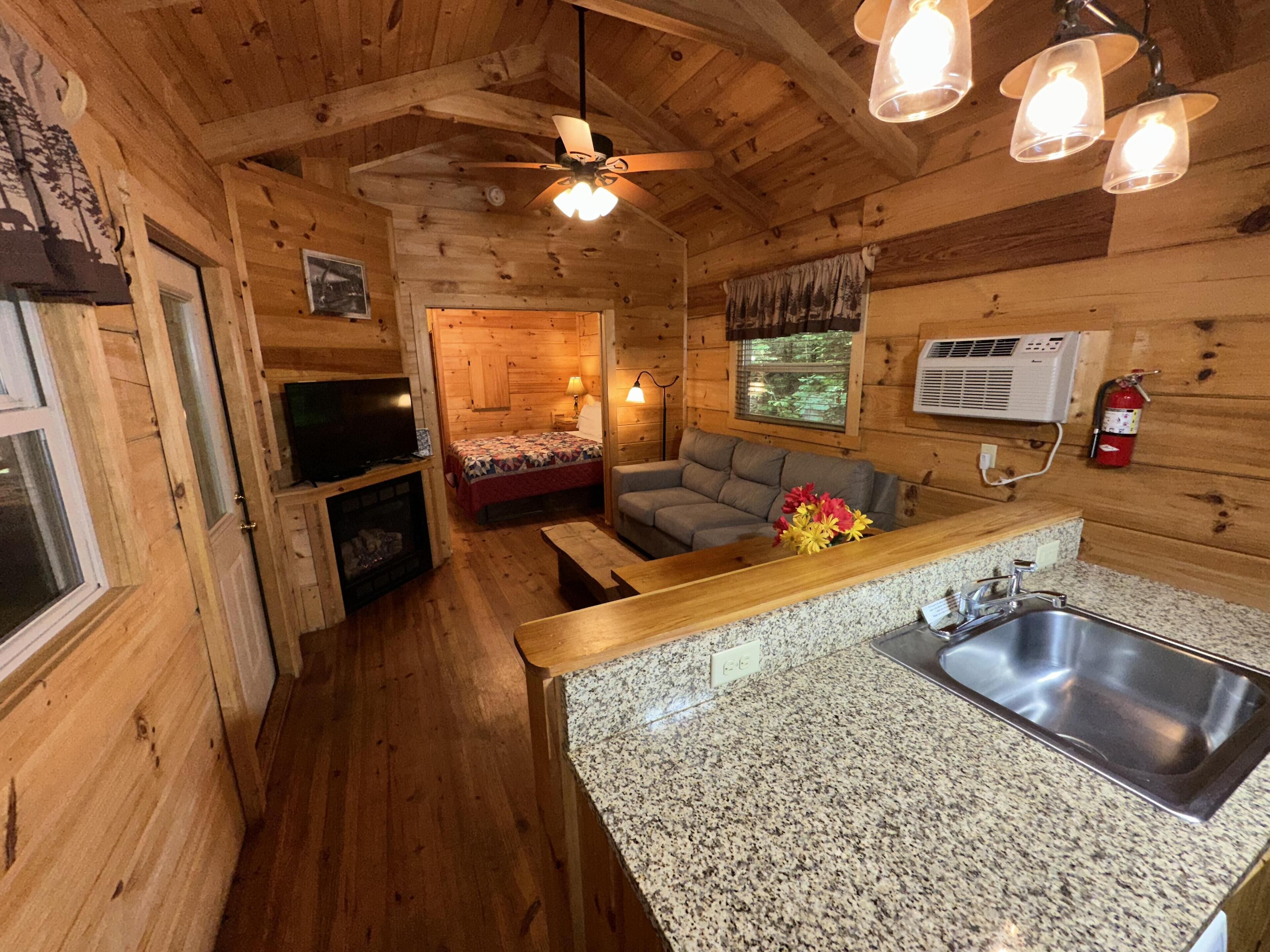 A wooden cabin interior with a kitchen, dining area, and a bedroom with a bed and window.
