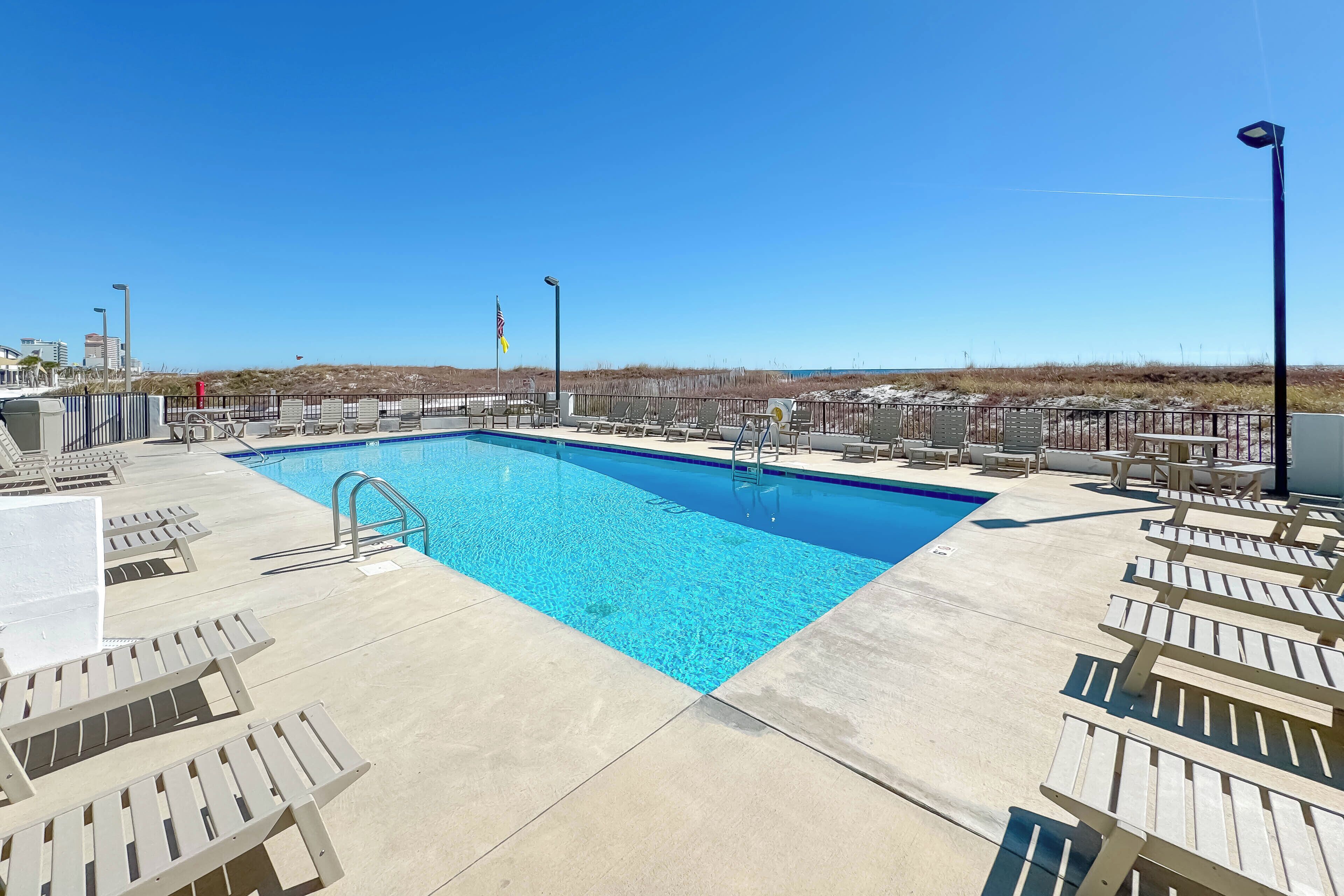 A rectangular swimming pool with a clear blue water surface, surrounded by lounge chairs and a paved deck.