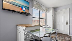Habitació moderna amb un televisor de temàtica marítima, una taula de menjador amb caires, una finestra amb vista a edificis i una porta blanca.