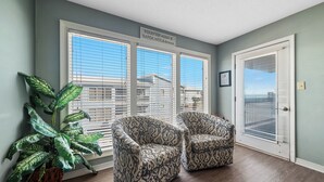 A room with two patterned armchairs, a large potted plant, and a view of a house through the windows. - PET FRIENDLY, TWO POOLS, LAGOON PIER, CONVENIENT TO THE BEACH, BEACHY DECOR (Gulf Shores)