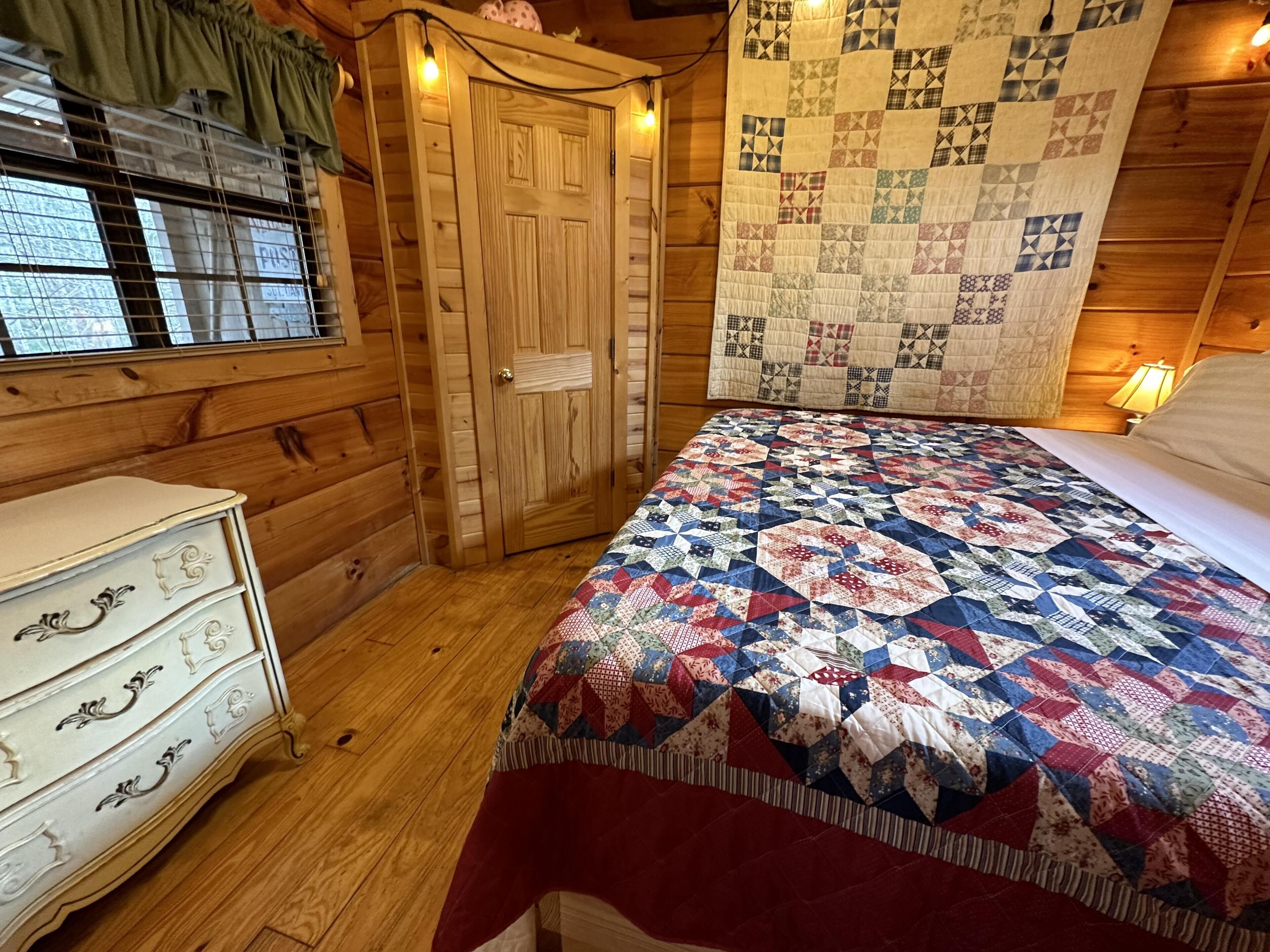 Una habitación acogedora con piso de madera, una cama con colcha colorida, un tocador con tiradores ornamentados, una puerta de madera y un tapiz acolchado en la pared.