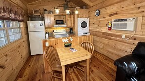 Een compact houten chalet met een keukenhoek, inclusief een koelkast, magnetron, fornuis en wasmachine. Er is een eettafel met stoelen en een klein decoratief plantje.