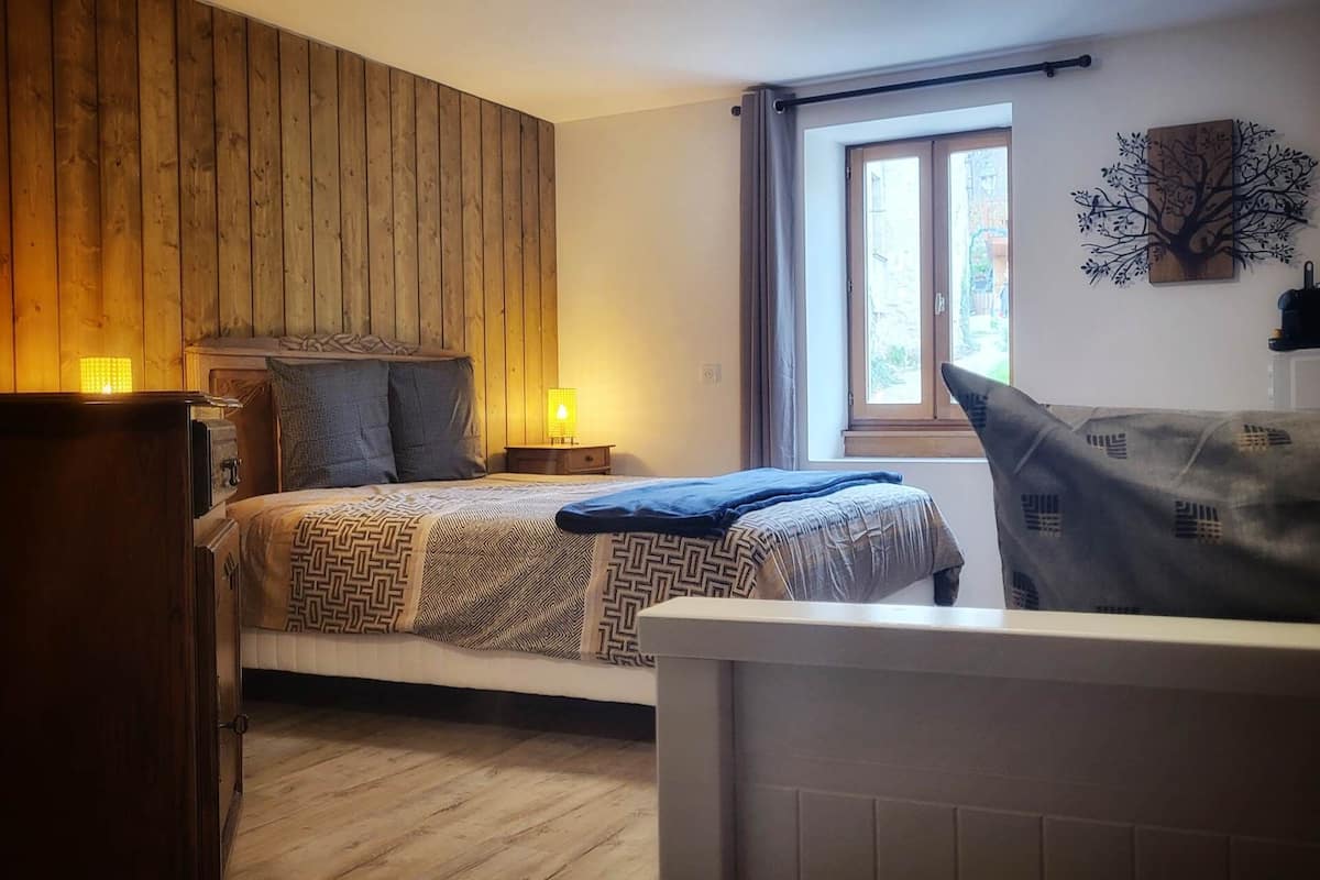 A bedroom with a wooden headboard, a bed with patterned bedding, a nightstand with a lamp, a window with curtains, and a wall-mounted tree decoration.