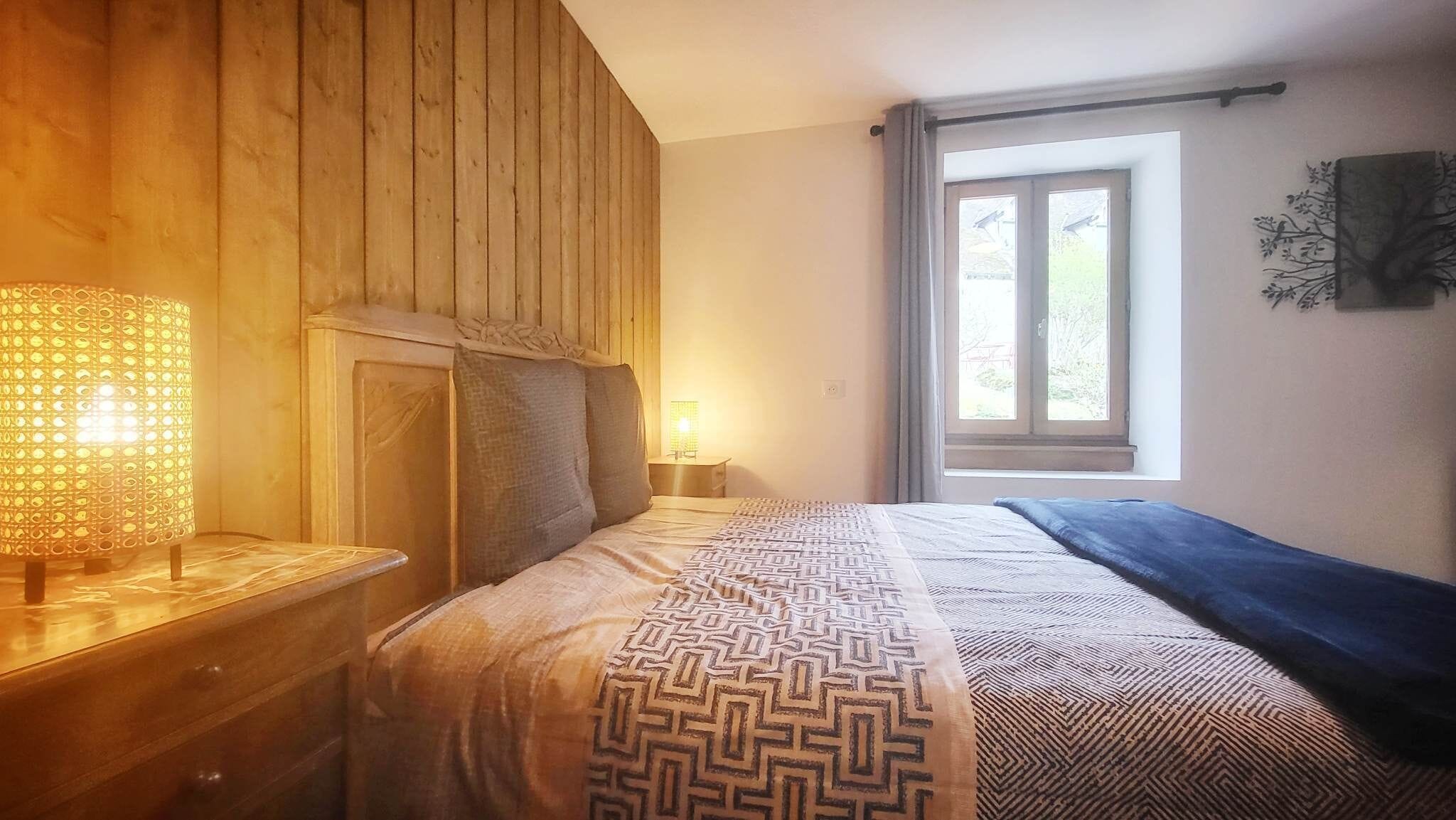 A bedroom with a wooden headboard, a patterned bedspread, a bedside lamp, and a window with curtains.