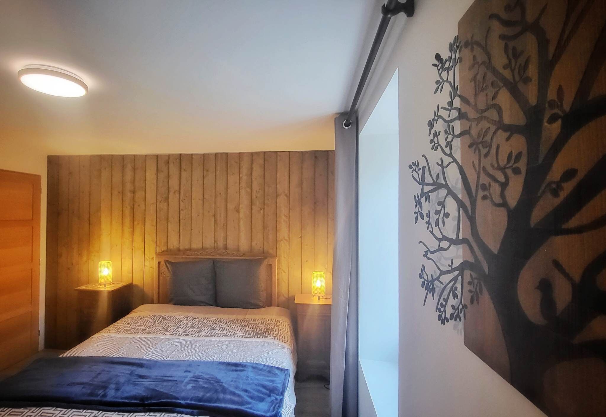 Un dormitorio con cabecera de madera, colcha estampada, dos lámparas de noche y un mural de un árbol en la pared.